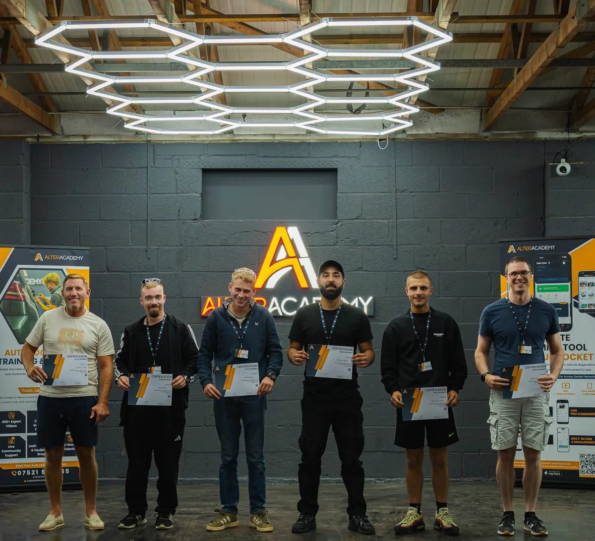 A class of six students holding their Alter Academy certificates under the hexagon lights and the Alter Academy sign