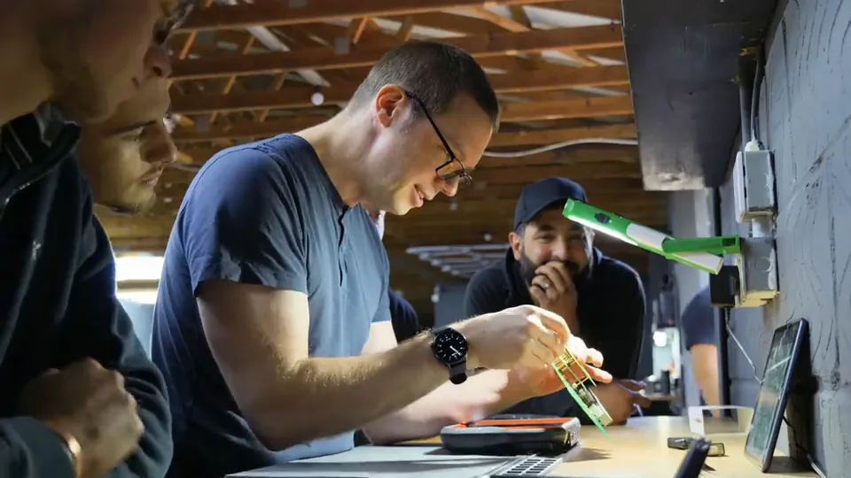 A trainee fits a green programming adapter at the bench while others look on