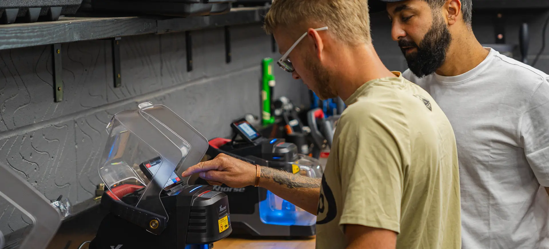 A trainee cuts a key on an Xhorse key cutting machine at the bench while a trainer watches