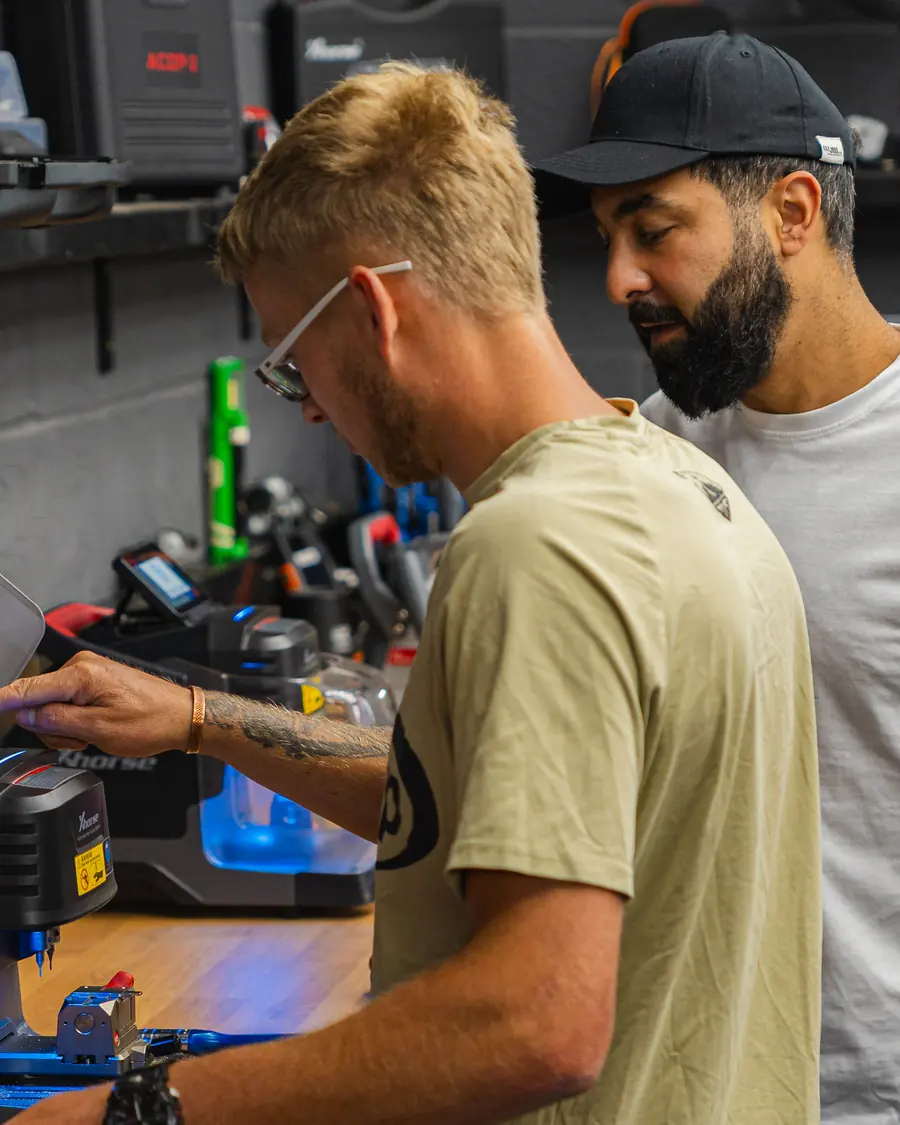 A trainee cuts a key on an Xhorse key cutting machine at the bench while a trainer watches
