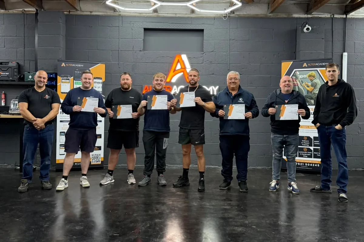 A class of students holding their Alter Academy certificates with their trainers, under the Alter Academy sign in the workshop