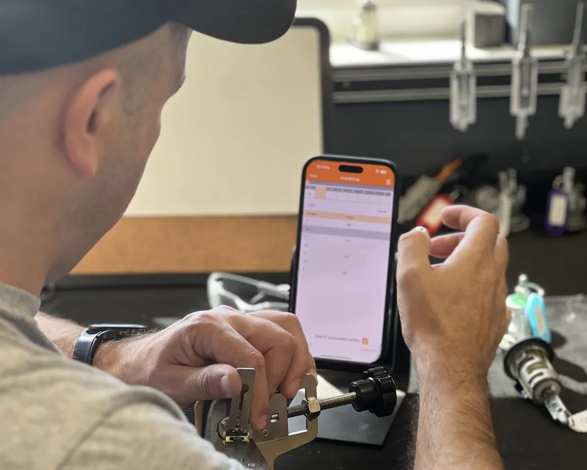 A trainee works a lock held in a bench jig with a pick tool, a phone on a stand beside him showing lock data